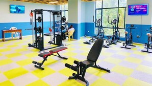 Une salle de sport équipée de matériel de musculation, de machines de cardio et d’un téléviseur affichant les prix.