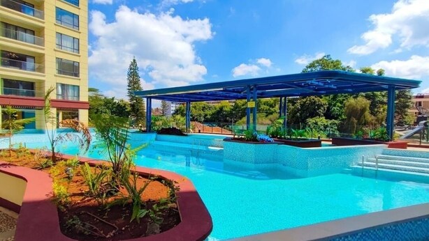 Une piscine dotée d’un auvent bleu, entourée de verdure et avec un bâtiment visible au loin.