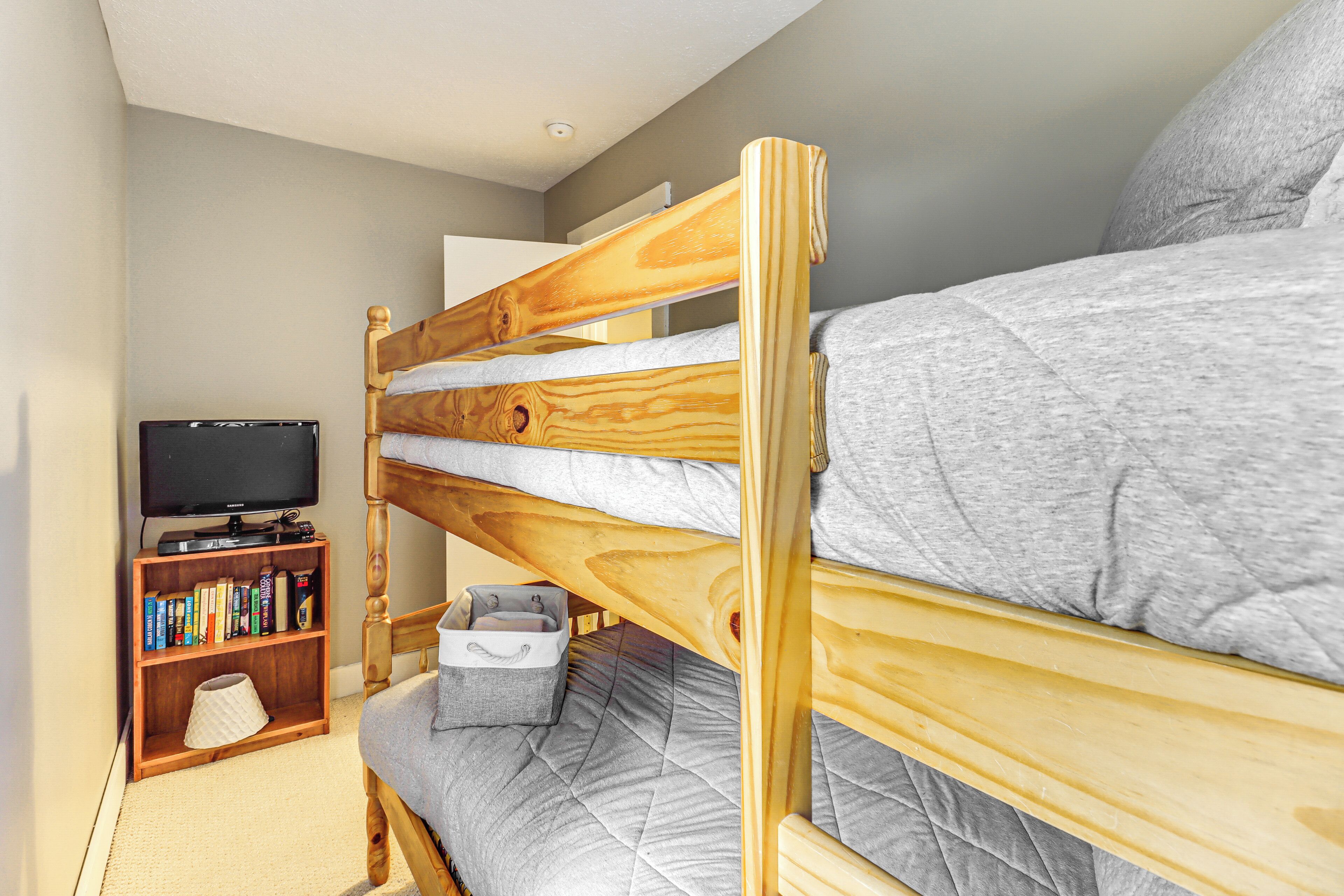 A bunk bed with a wooden headboard, a grey mattress, and a small bedside table with a lamp. A bookshelf with books and a flat-screen TV is visible in the background.