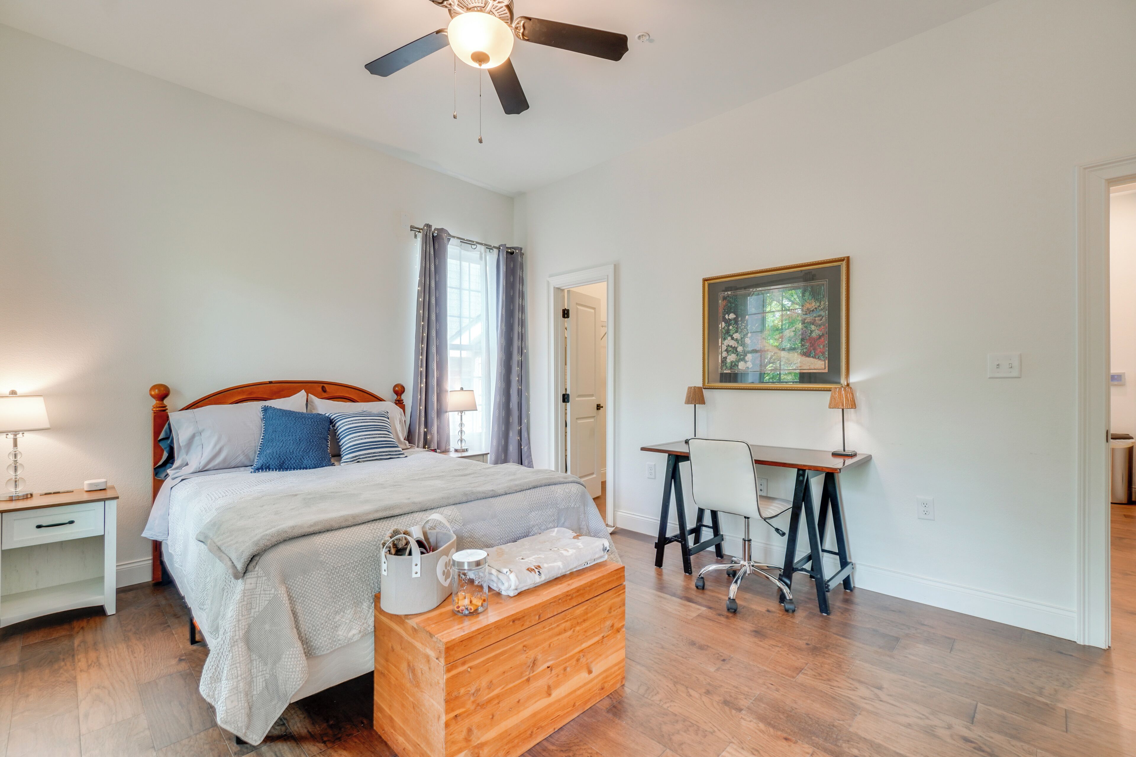 A bedroom with a bed, a desk, a chair, a ceiling fan, and a painting on the wall.