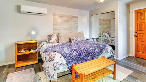 Une chambre à coucher comprenant un lit, une table de chevet, un banc, un climatiseur, un ventilateur de plafond et une porte.