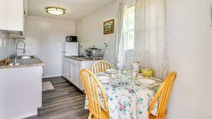 Une cuisine compacte avec des meubles blancs, un four à micro-ondes et un coin repas agrémenté d’une nappe à motifs floraux et de chaises en bois.