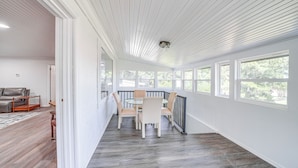 A spacious room with a dining area featuring a round table and chairs, large windows, and a view of greenery outside.