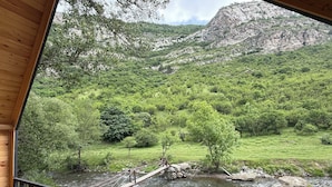 A wooden deck overlooking a river with a bridge, surrounded by lush greenery and mountains.