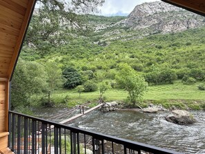 A wooden deck overlooking a river with a bridge, surrounded by lush greenery and mountains.