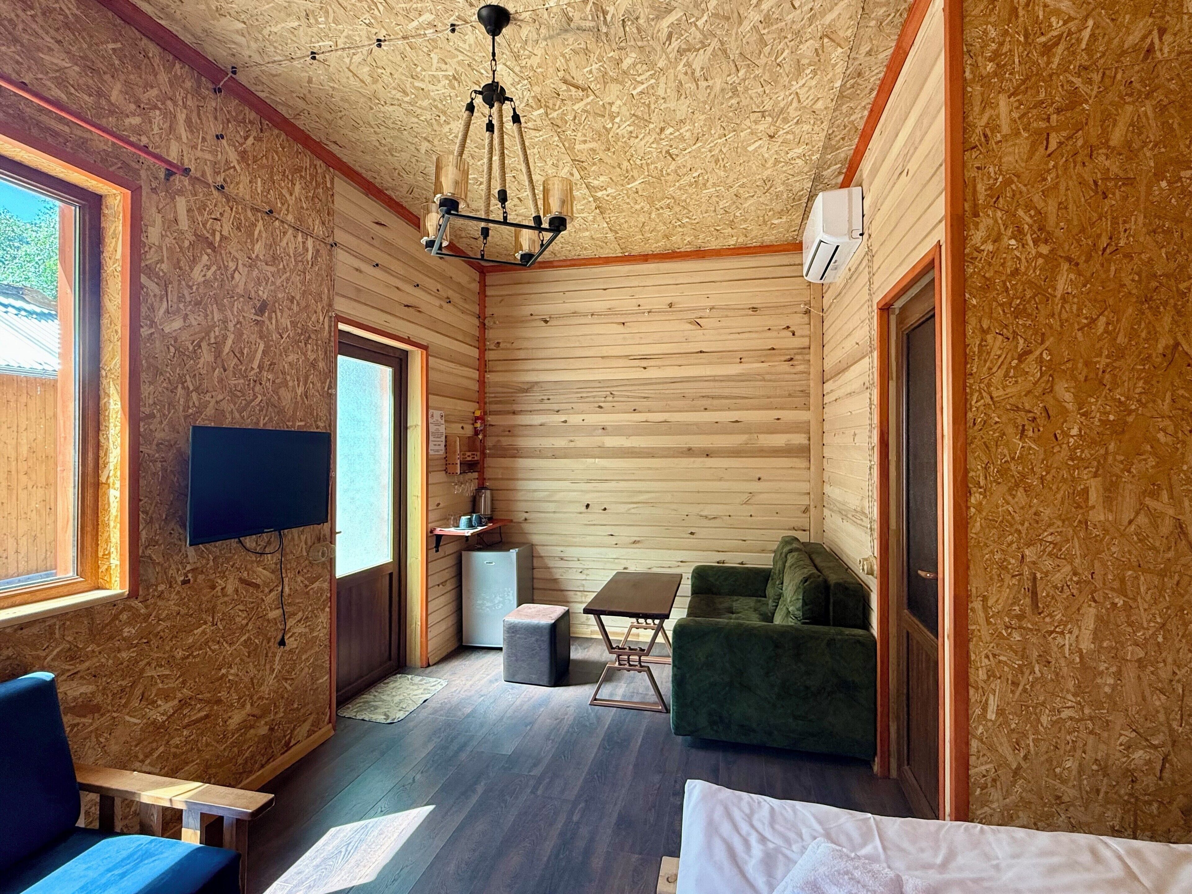 Interior de una cabaña con detalles de madera, un televisor montado en la pared, un sofá verde, una mesita y un accesorio de iluminación en el techo.
