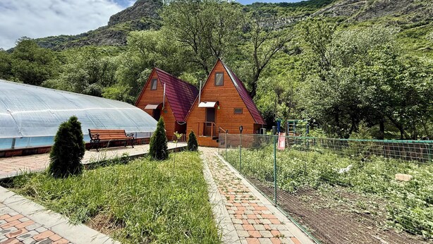 A wooden cabin with a red roof, surrounded by greenery and a greenhouse.