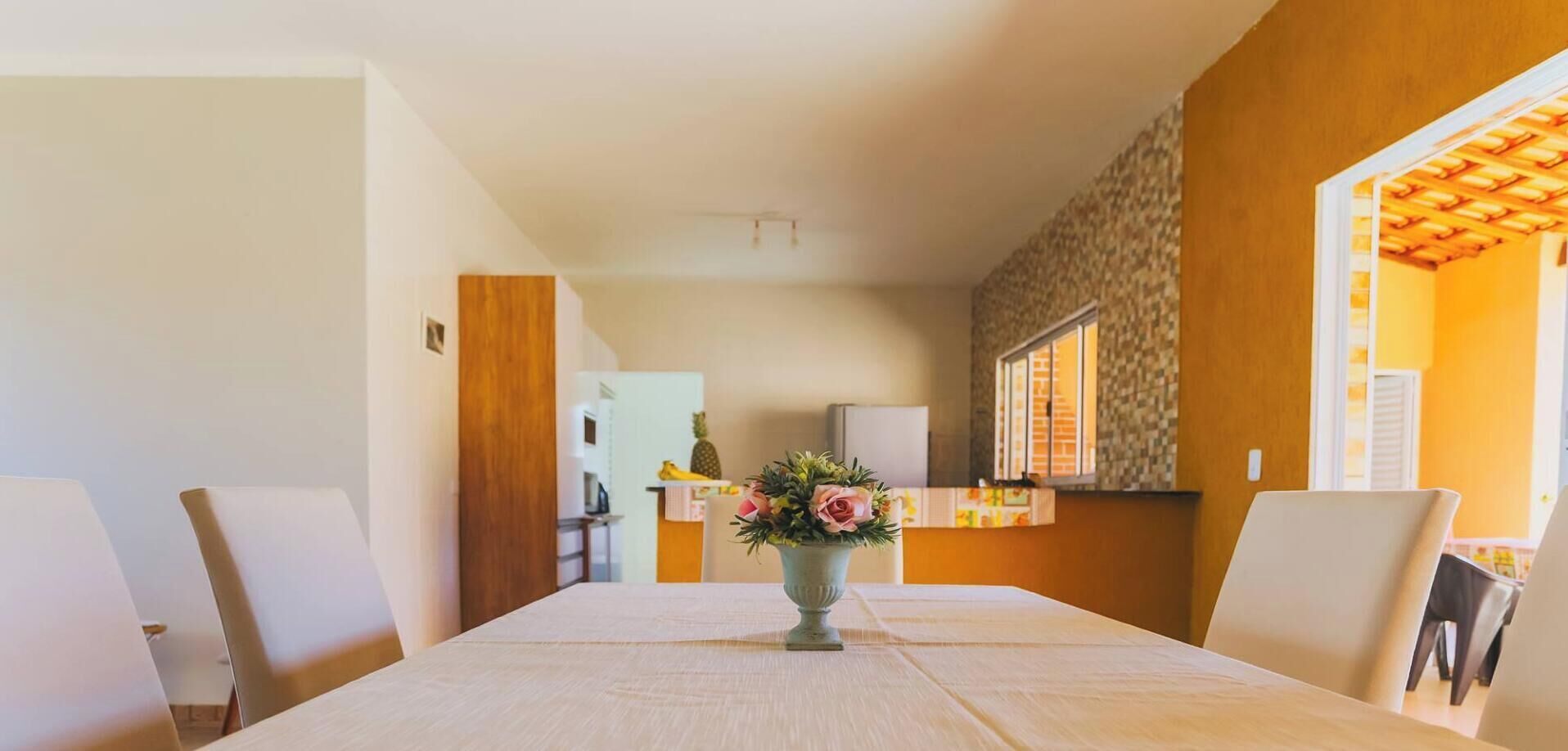 A dining area with a table set for four, a vase with flowers, and a kitchen in the background.