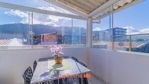 A balcony with a table and chairs, a potted plant, and a view of a mountainous landscape and buildings. - Apartamento em Ubatuba com Churrasqueira (Ubatuba)