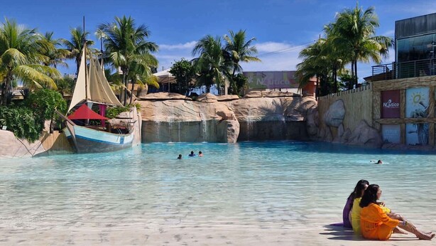 A pool area with a sailboat, palm trees, and a rocky backdrop.