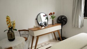 Un dormitorio con cama, mesita de noche con espejo, pequeño escritorio con un florero, una silla y un ventilador de pie.