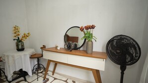Una habitación pequeña y ordenada con un escritorio de madera, un espejo, un florero con flores, un ventilador y una pequeña mesa con mantel de encaje.