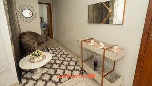 Un espacio habitable pequeño con un sofá, una mesita redonda con un centro de mesa floral, un espejo, un adorno en la pared, una vitrina con tazas y vasos, y un cuadro en la pared.