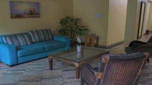 Uma sala de estar com um sofá azul, uma mesa de centro de vidro, cadeiras de vime, uma planta em vaso e um quadro na parede.
