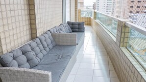 Varanda com sofá cinzento, cadeiras de vime, paredes azulejadas e vista para edifícios.