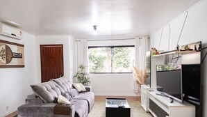 Un salon moderne avec un canapé gris, une table basse, un téléviseur posé sur un meuble et une fenêtre donnant sur des arbres.