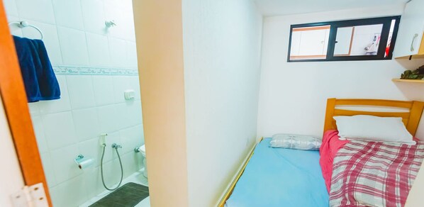A small, compact room with a bed, a window, and a shower area. - Apartamento com Vista para o Mar em Guarapari (Guarapari)