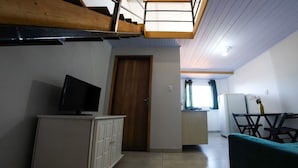 Un espacio habitable compacto con un televisor sobre un mueble, una zona de comedor con sillas y una cocina al fondo.