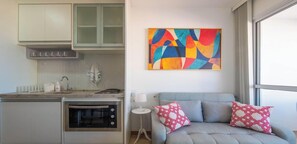 Uma cozinha moderna com forno embutido, área de jantar com sofá cinza e almofadas estampadas, e um quadro abstrato colorido na parede.