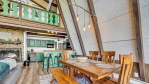A loft-style dining area with a wooden table and chairs, a stone fireplace, and a kitchen with green cabinets. - Press Pause | Hot Tub + Epic Mountain Views (Beech Mountain)