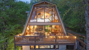 A two-story wooden cabin with a balcony, surrounded by trees. - Press Pause | Hot Tub + Epic Mountain Views (Beech Mountain)