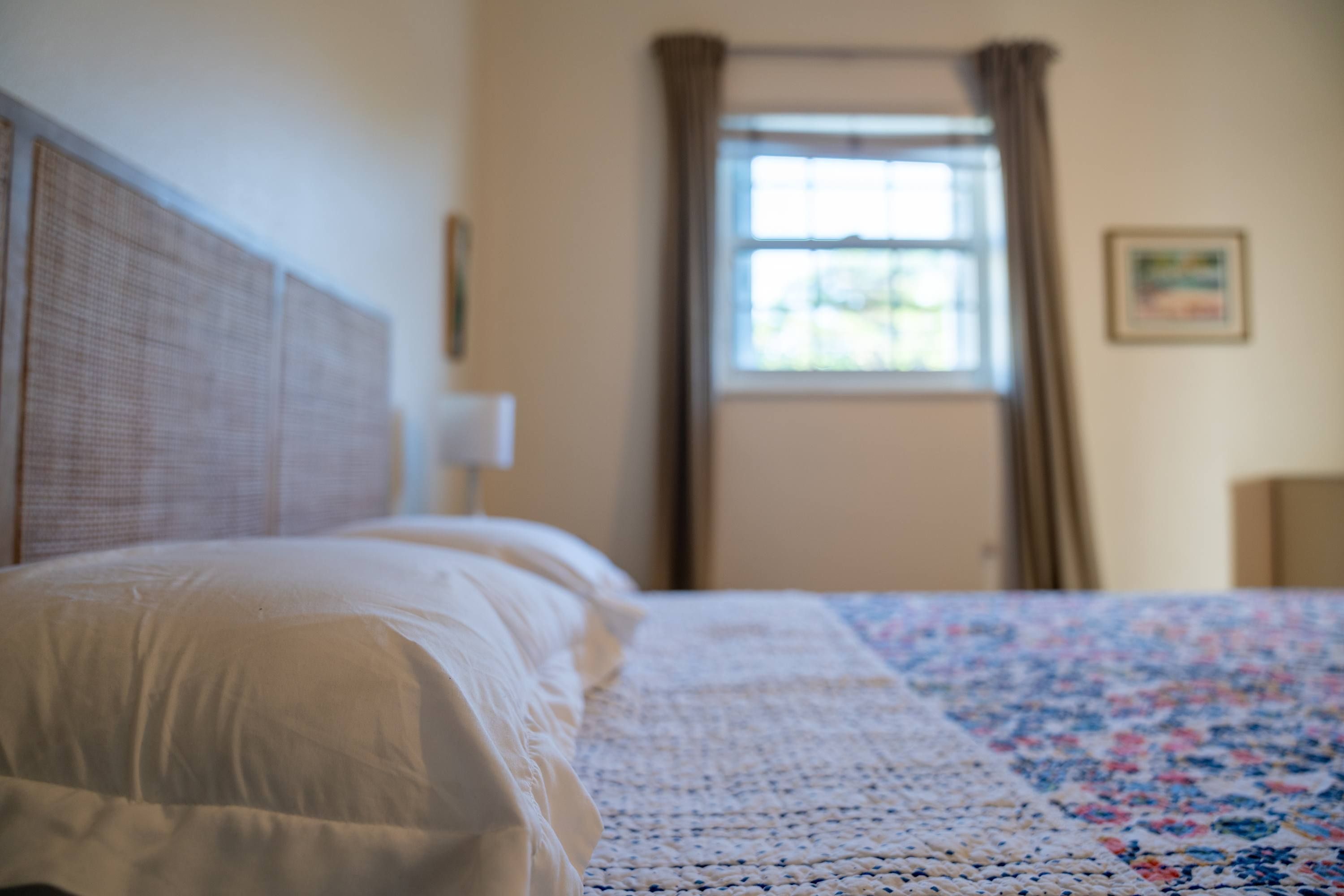 Una cama con cabecera blanca, una colcha estampada con motivos florales y una ventana con cortinas.