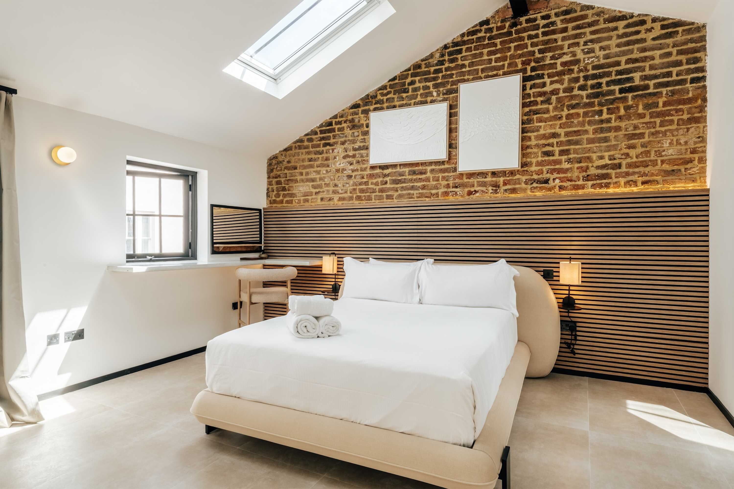 A modern bedroom with a brick wall, a skylight, a bed with white linens, and a bedside table with a lamp.