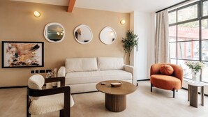 A modern living room with a beige sofa, a round coffee table, a wooden chair, and an orange armchair. There are three wall-mounted mirrors and a framed abstract painting. - The Primrose Mews in Greater London (London)