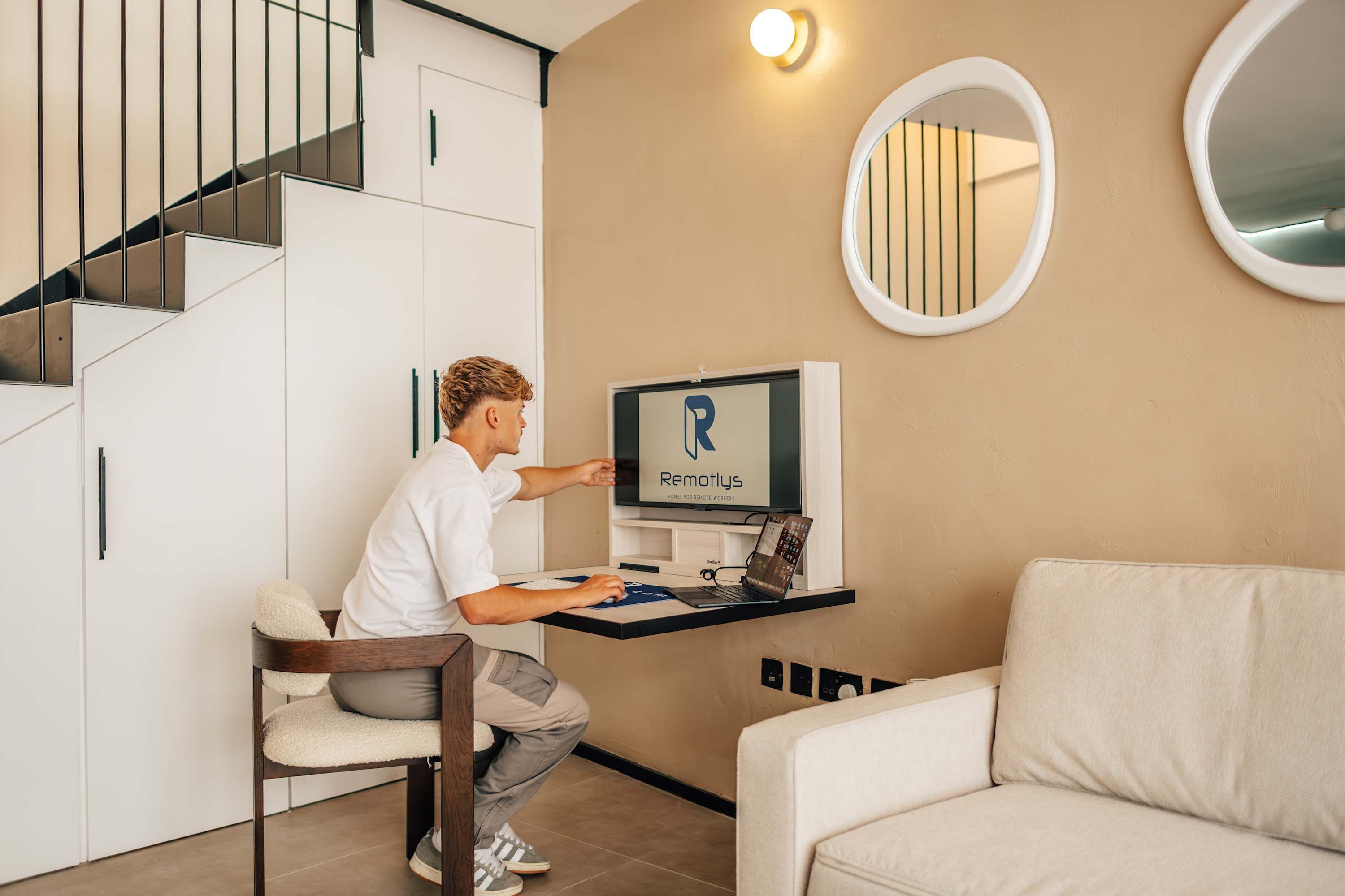 Iemand gebruikt een laptop aan een bureau, met een Remollys bord, een bank en een trap in een modern interieur.