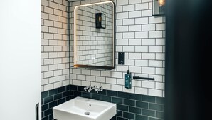 A modern bathroom with a white sink, black and white tiled walls, a mirror with a light, and a black towel dispenser. - The Primrose Mews in Greater London (London)