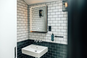 A modern bathroom with a white sink, black and white tiled walls, a mirror with a light, and a black towel dispenser. - The Primrose Mews in Greater London (London)