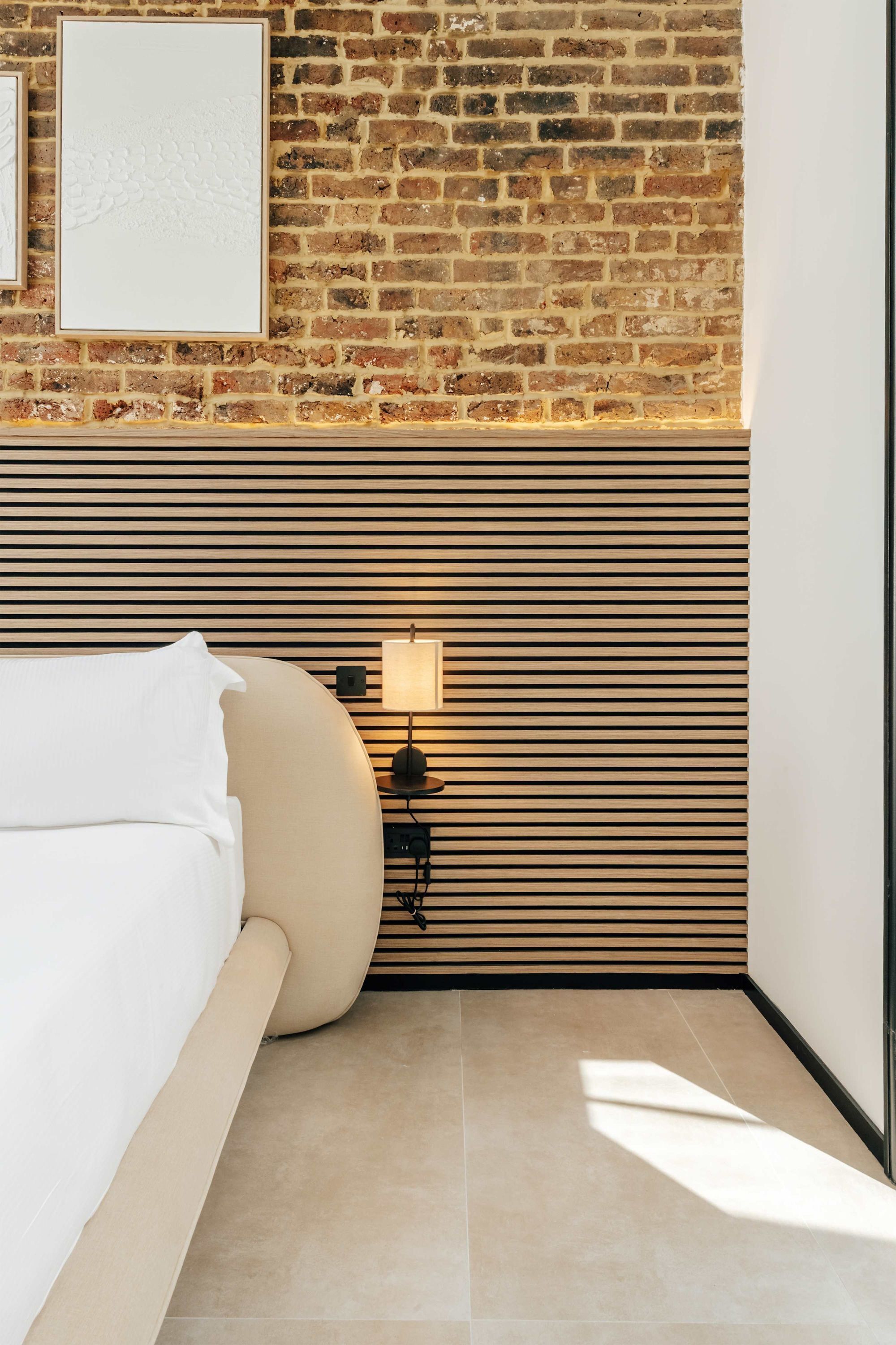 A modern bedroom with a brick wall, a slatted panel design, a bed with white bedding, and a bedside table with a lamp.