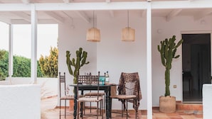 Un patio couvert avec une table et des chaises, deux lampes suspendues en osier et deux grands cactus.