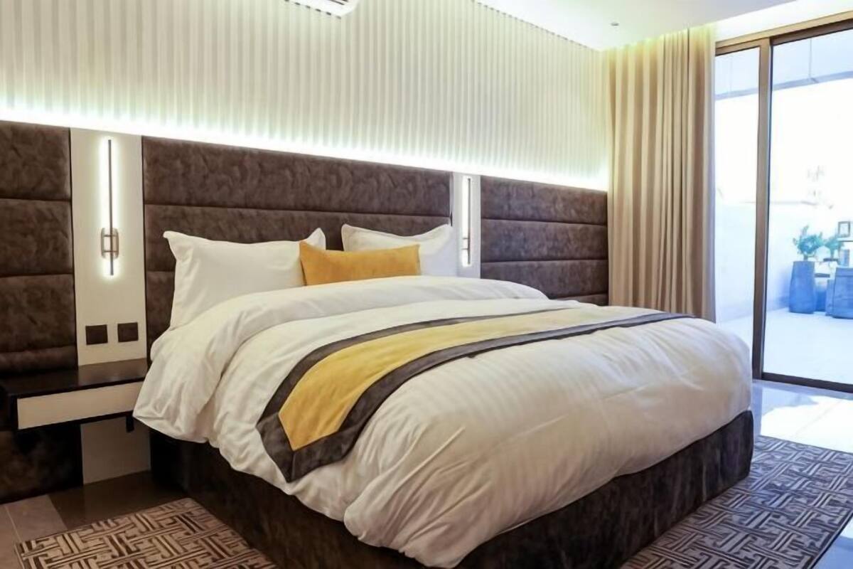 Quarto de hotel moderno com uma cama grande, cabeceira com iluminação integrada, mesa de cabeceira e vista através de uma porta de vidro.