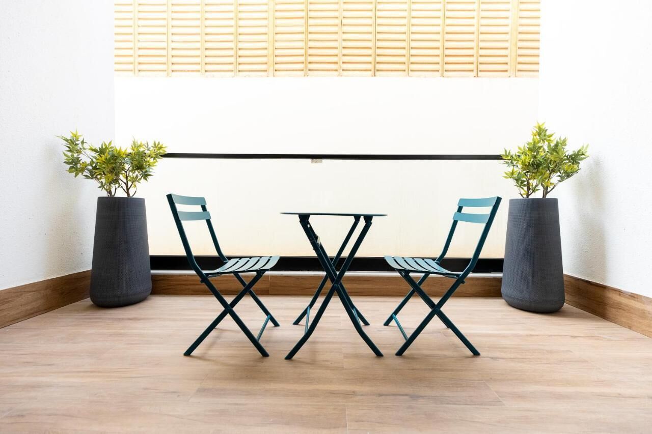 A small, modern room with a wooden floor, a white wall, and a window with wooden blinds. There is a small table with two chairs and two potted plants.