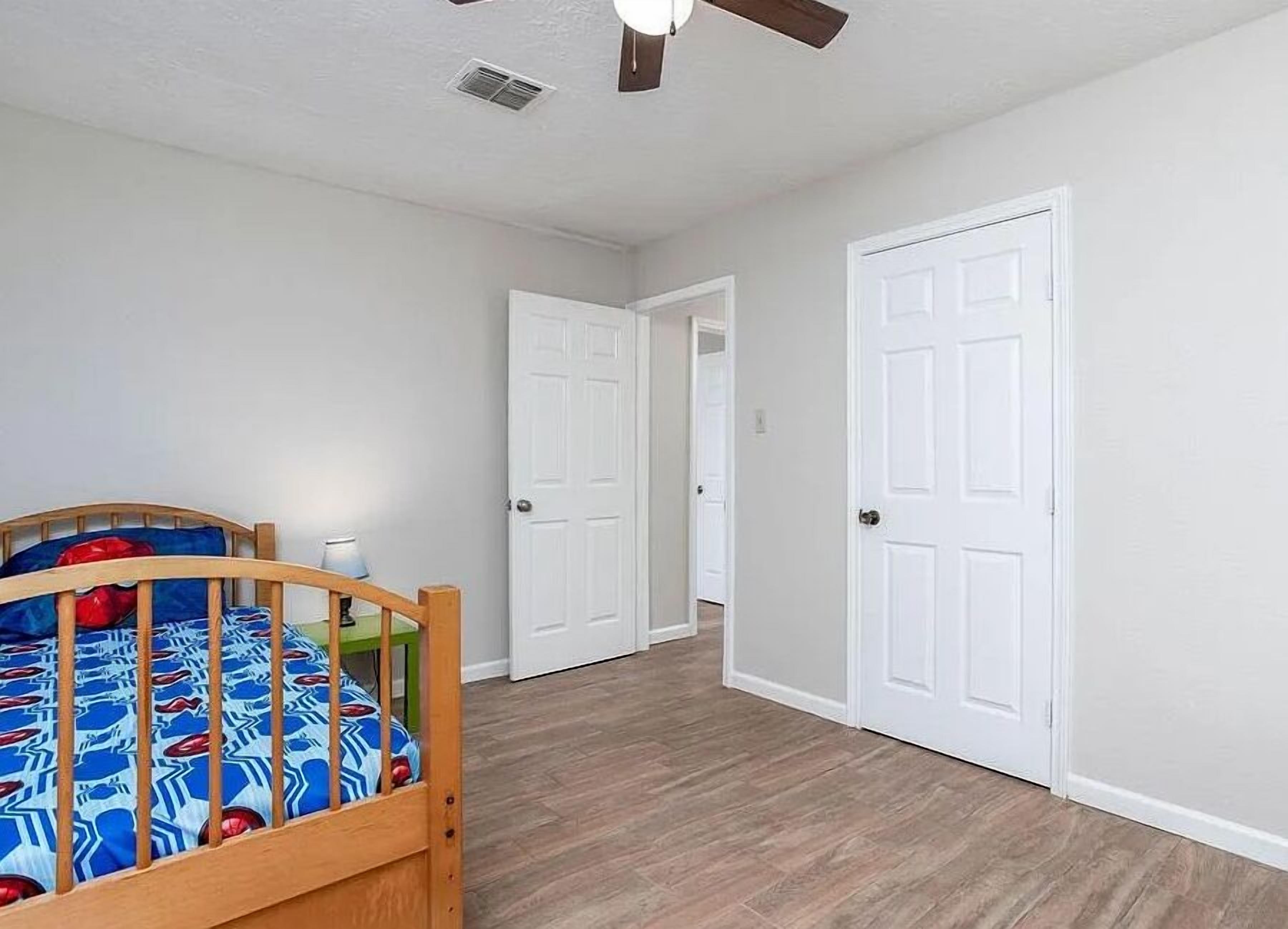 A bedroom with a wooden bed, white walls, and a wooden floor. There are two white doors and a ceiling fan.