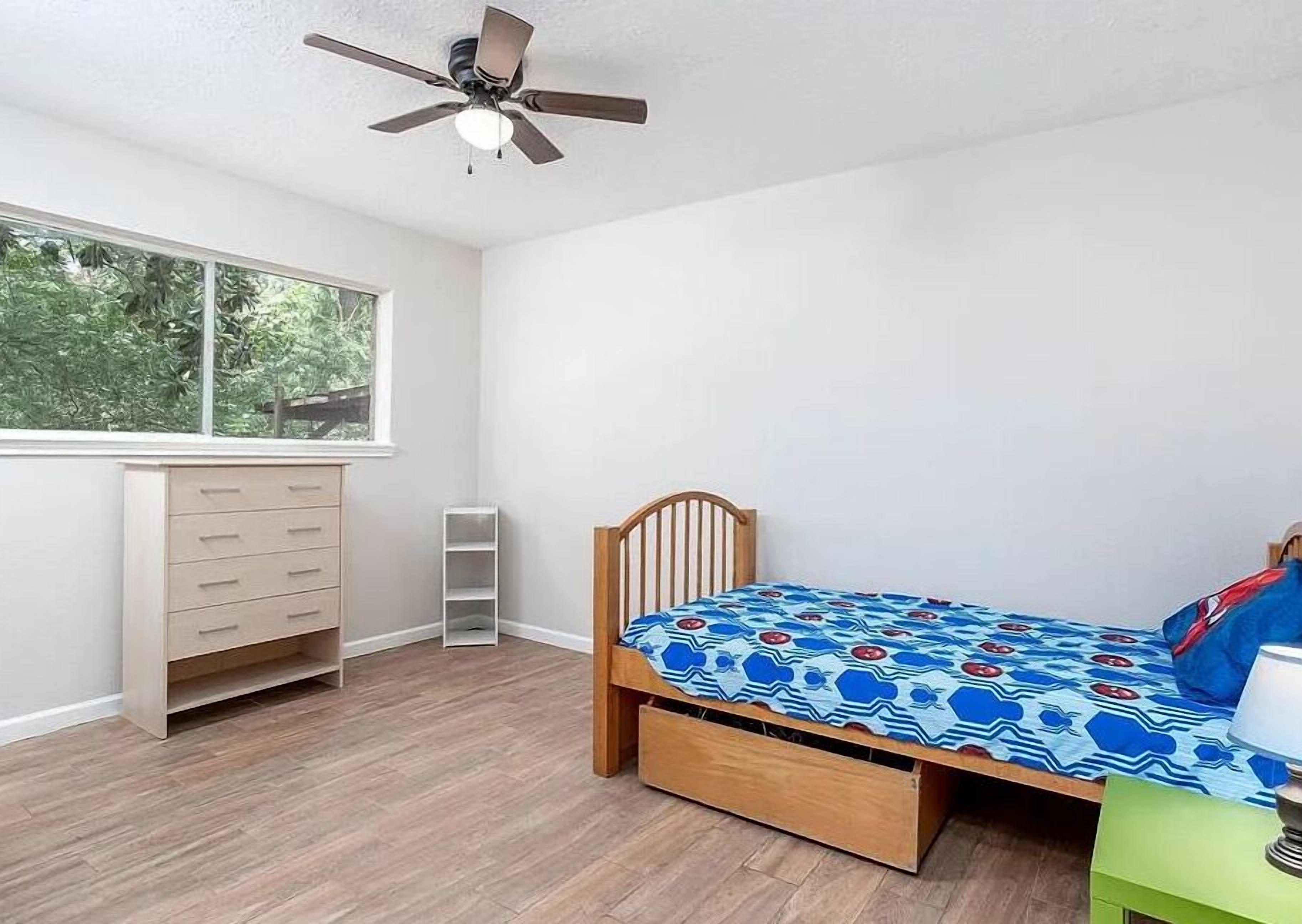 A bedroom with a bed, a desk, a chair, a ceiling fan, and a window.