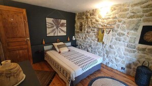A bedroom with a stone wall, a bed with patterned bedding, a wooden door, and a small shelf with decorative items. - Family villa in the luberon regional park with swimming pool for 6 people (Puget)