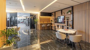 A modern interior with a glass partition leading to a gym area, a round table with chairs, and wooden paneling with shelves and a TV. - StayShort - Stylish DIFC Apartment Sleeps 4 with Pool & Gym (DIFC)
