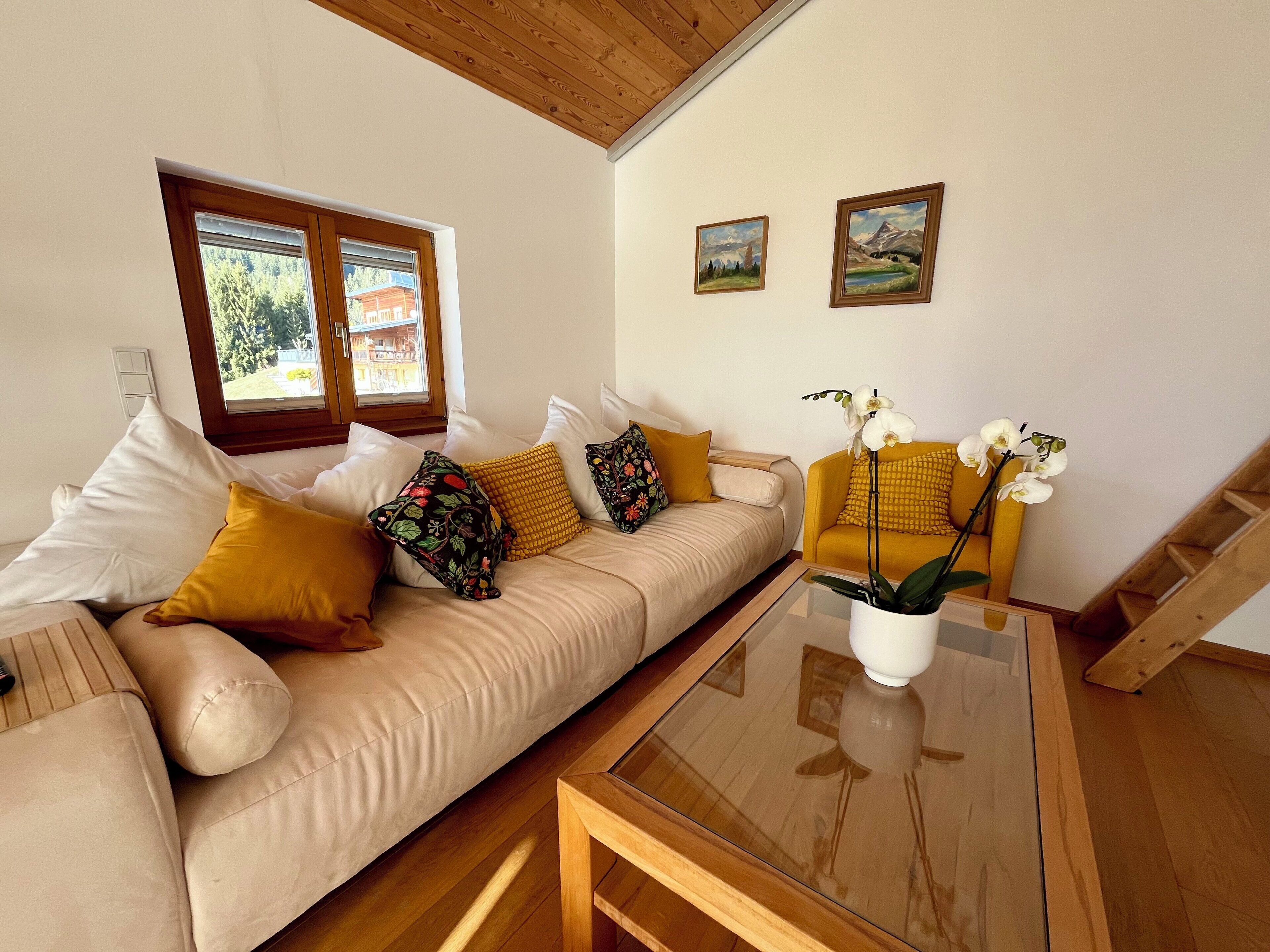 Ein modernes Wohnzimmer mit einem beigen Sofa, gelb-blumigen Kissen, einem Glastisch und zwei gerahmten Landschaftsbildern an der Wand.