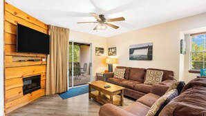 A living room with a flat-screen TV, wooden wall paneling, a ceiling fan, a sofa set, and a fireplace. - Cozy Timbers - Charming Condo for 6 with Resort-style Amenities (Branson)