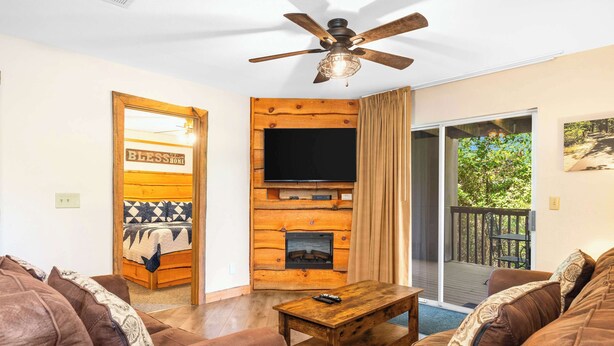 A cozy living room with a fireplace, a ceiling fan, and a large window overlooking a deck. - Cozy Timbers - Charming Condo for 6 with Resort-style Amenities (Branson)