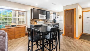 A modern kitchen with dark cabinets, a central island, and a dining area with chairs. - Cozy Timbers - Charming Condo for 6 with Resort-style Amenities (Branson)