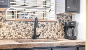 A kitchen with a stone backsplash, a coffee maker, and a sign that reads "friends and family gather." - Cozy Timbers - Charming Condo for 6 with Resort-style Amenities (Branson)