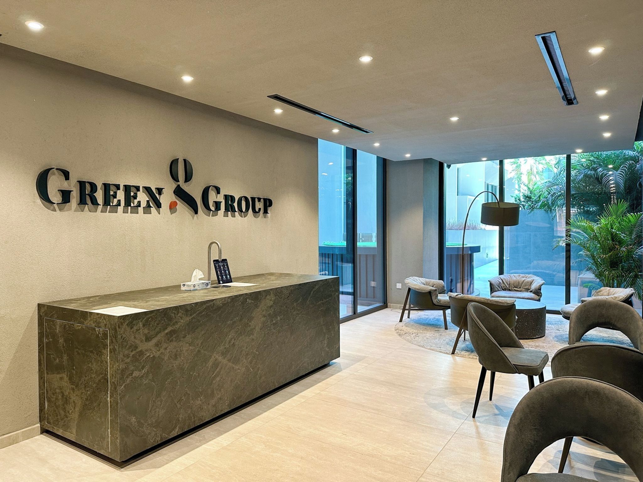 Una recepción con encimera de mármol, un letrero en la pared que dice 'GREEN GROUP', y una zona de estar con sillas y una mesa redonda.