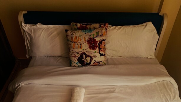 Un lit bien fait, avec des draps blancs, un coussin à motifs floraux et une serviette enroulée.