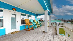 A covered porch with wooden flooring, a row of colorful chairs, and a view of the ocean and buildings in the background. - Puddin Place: 3 BR, 2.5 BA House in Crystal Beach, Sleeps 10 (Bolivar Peninsula)