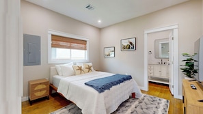 Un dormitorio con cama, mesita de noche, armario, ventana con persianas y un baño con espejo y lavabo.
