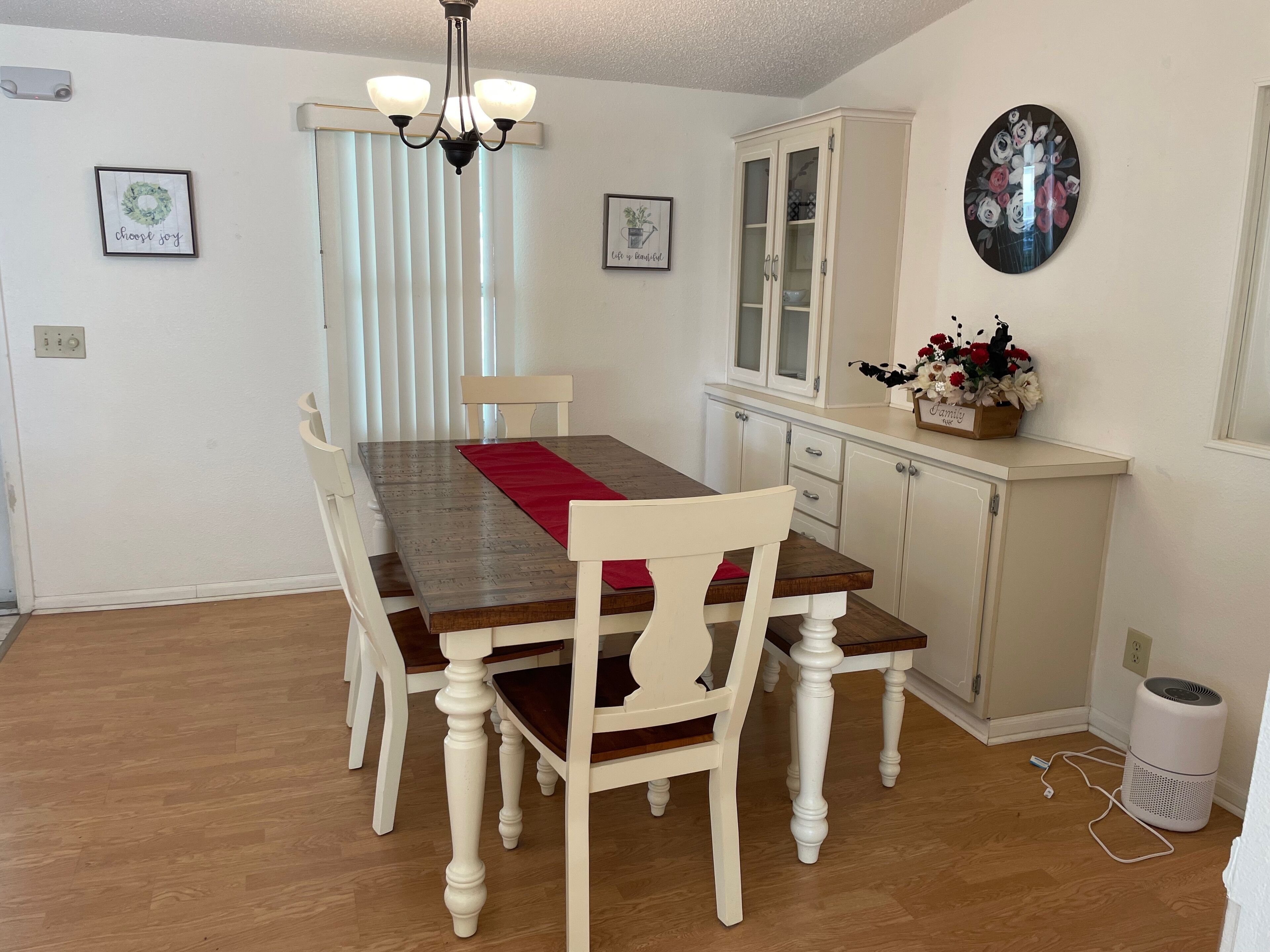 Un coin repas comprenant une table en bois, des chaises blanches, un lustre, un buffet et un tableau accroché au mur.