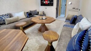 A modern living room with a wooden coffee table, a round side table, and a sofa set. A large TV mounted on the wall displays the Netflix logo.